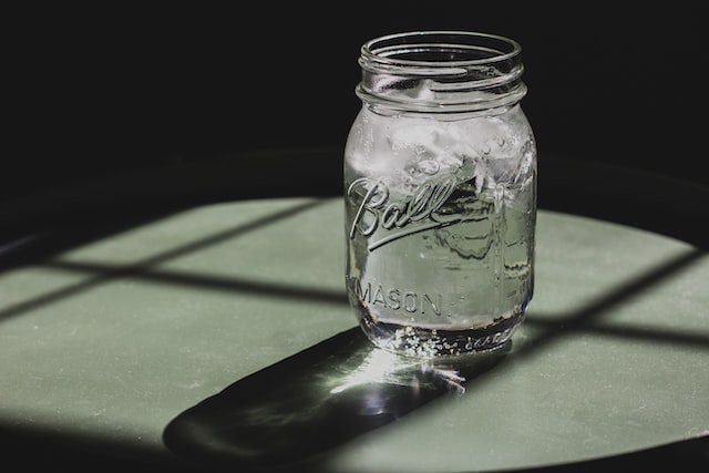 A mason jar filled with ice water placed on a table, casting a shadow in the sunlight.