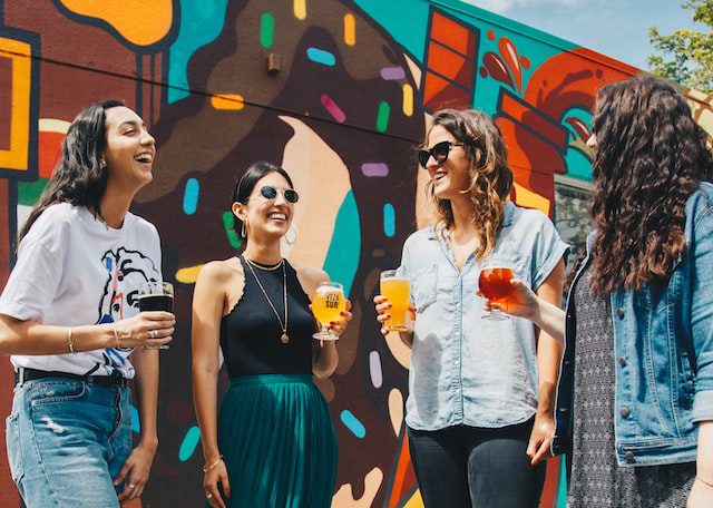 Four women sharing a laugh while holding drinks in broad daylight - a perfect depiction of a warm and welcoming atmosphere in a shared living space. Discover the 10 proven tips to break the ice with your new roommates and build lasting friendships in your new home.