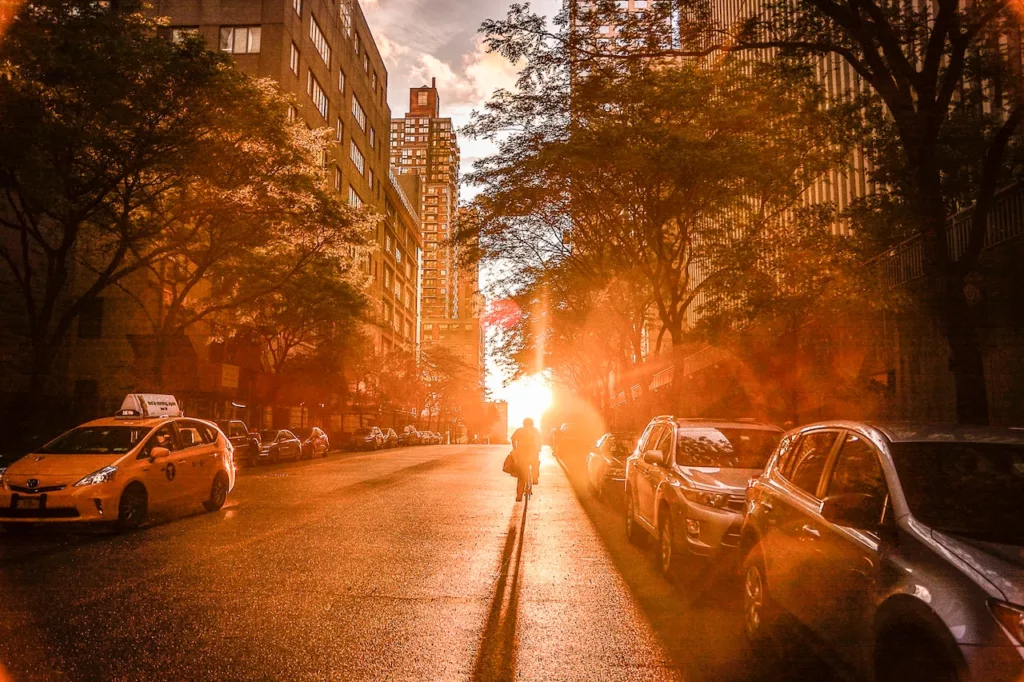 A picturesque sunny day in New York City, with bustling sidewalks filled with cars and a person riding a bicycle towards the sunset, representing one of the best neighborhoods for young professionals to reside in.