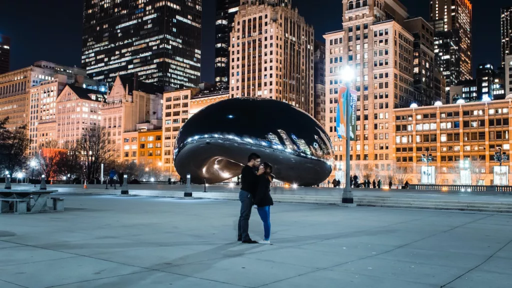 A couple standing in front of the illuminated Cloud Gate sculpture in Chicago, Illinois, sharing a romantic kiss at night, as they uncover the city's hidden romantic gems.