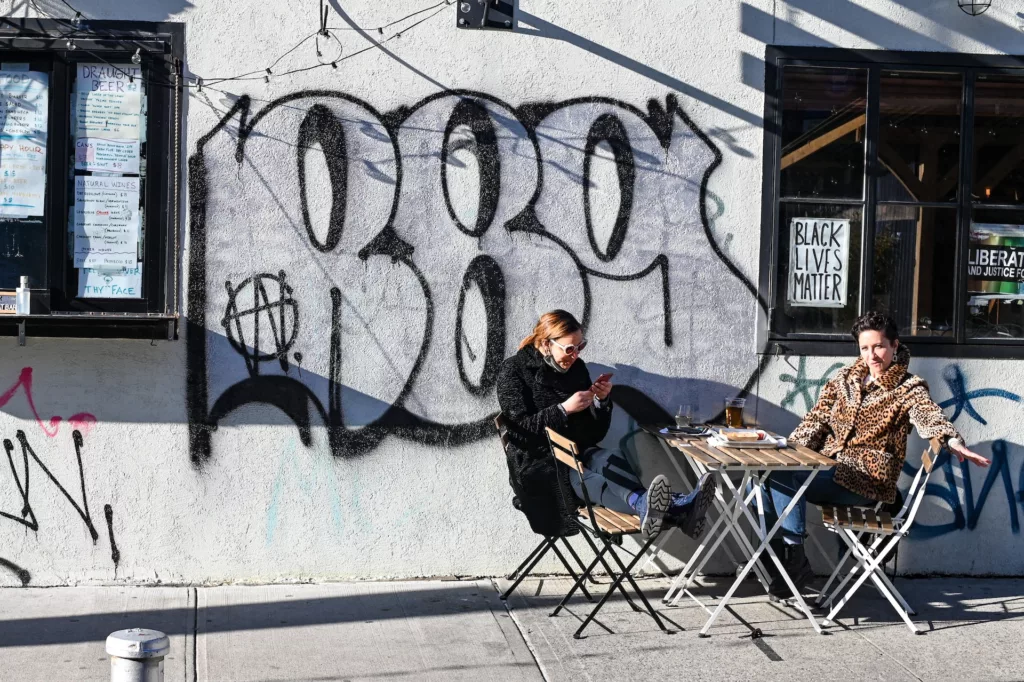 Man sitting on a folding chair in front of graffiti-covered wall in Brooklyn, a representation of the borough's urban and artistic culture, ideal for Moving To Brooklyn Essentials: Your Guide To Brooklyn Life article.