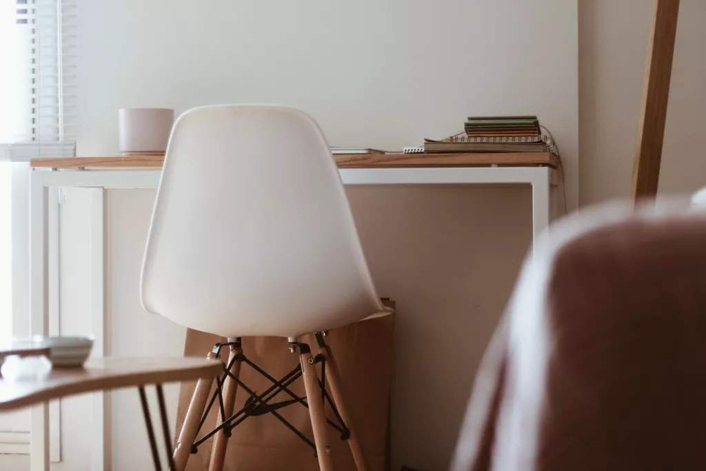 A minimalist home office setup featuring a white chair next to a wooden desk with a stack of books and a small cup. A window with white blinds illuminates the serene workspace. Ideal for articles discussing rental spaces and interior design.