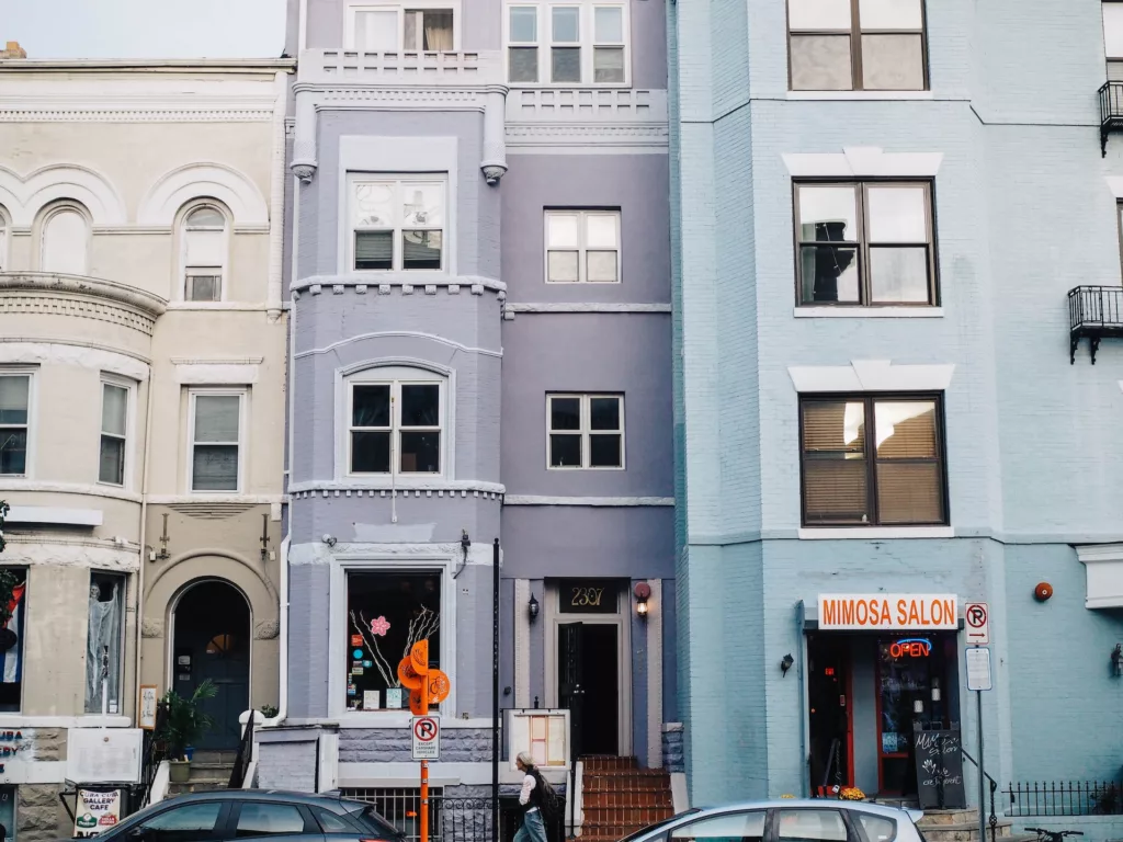 Row of colorful townhouses with architectural details, including a lavender-painted building and a light blue one. The ground floor of the blue building has a storefront labeled 'Mimosa Salon' with an 'Open' sign. A few cars are parked in front, and a pedestrian is visible on the sidewalk.