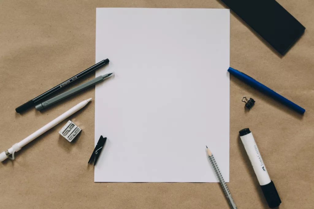 A flatlay of stationary items on a beige background, including a blank white paper, black and white pens, a pencil, eraser, pencil sharpener, and a blue pencil cap.