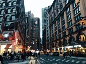 Evening view of East Village in Manhattan with bustling pedestrians crossing the street, illuminated store signs, and a blend of historic and modern buildings lining the road.