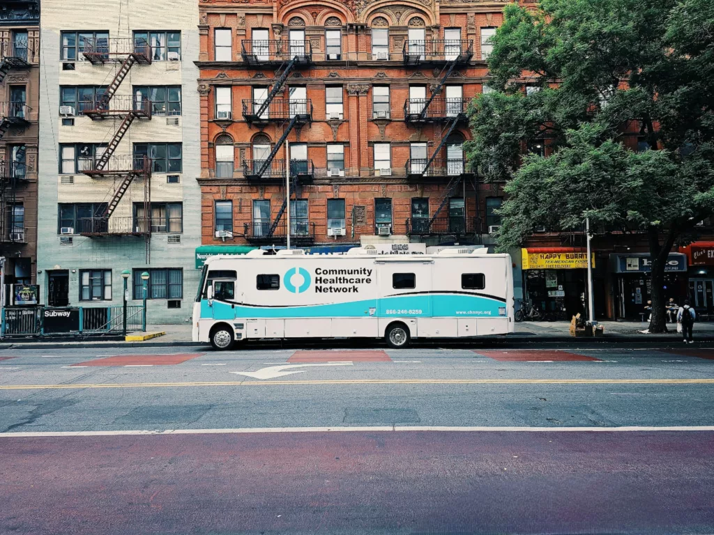 Street view of East Village, Manhattan, showcasing historic brick buildings, a Community Healthcare Network van, and local businesses, with pedestrians walking on the sidewalk.