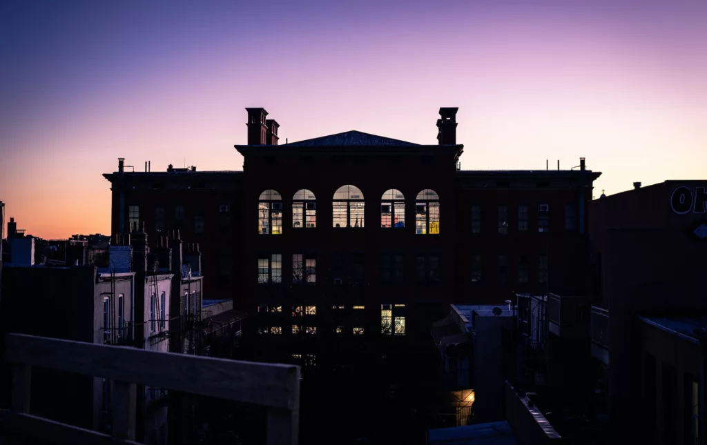 Evening descends on Bushwick, showcasing the silhouette of a distinguished red-brick building with large windows that glow against the twilight sky. In the foreground, the darkened outlines of residential buildings and fire escapes hint at the urban density, while the fading light paints a serene gradient from warm purple to deep blue above.