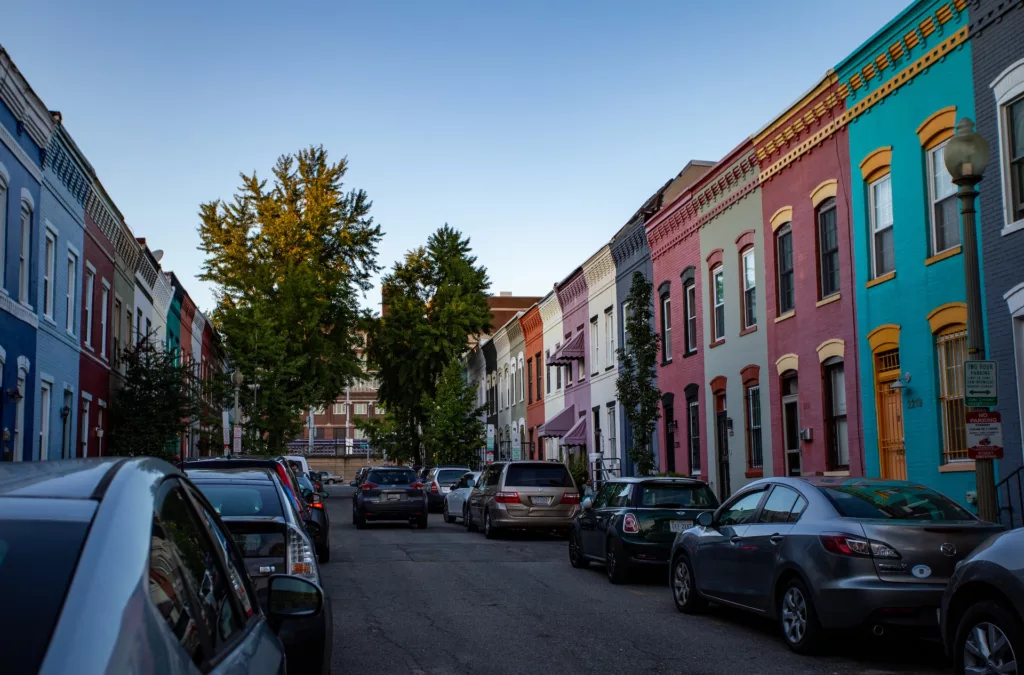 A street view of a row of colorful townhouses in an urban residential area with cars parked on both sides of the street, suggesting a densely populated neighborhood. The architecture indicates a mix of historical charm with vibrant modern colors, typical of a metropolitan area such as Washington D.C. The image is taken during the day under clear skies.