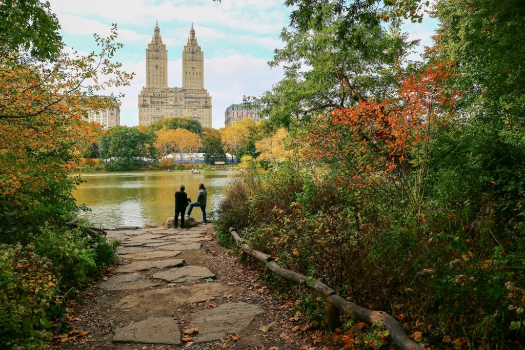 Two people standing by the edge of a tranquil pond in Central Park, New York City, with the iconic San Remo apartment buildings rising majestically in the background amidst lush autumn foliage.