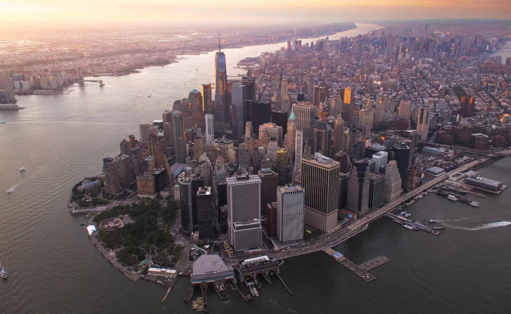 Aerial view of Lower Manhattan at sunset, with the Hudson River on the left and the orange hues of the setting sun reflecting off the skyscrapers, highlighting the dense urban landscape of New York City.