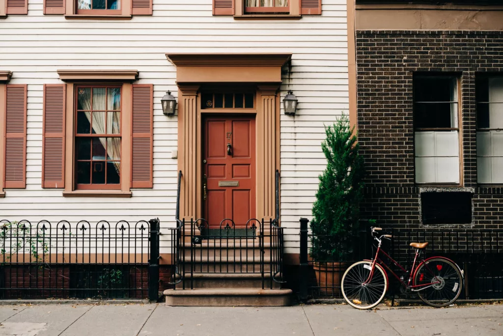 This image depicts a quaint urban scene, featuring the facade of a residential building. The building's exterior is a combination of cream siding with dark red shutters on the windows, and a brick section painted in a contrasting darker color. A robust red door is centered in the frame, providing a warm and inviting entrance, accented by a small overhead portico and flanked by classic-style lamps. A simple black metal fence with decorative elements borders the building, and a solitary bicycle with a red frame and brown saddle is parked against the fence on the sidewalk, adding a touch of urban lifestyle to the scene. The overall impression is of a peaceful, charming street view in a city, perhaps in the morning or late afternoon, given the soft lighting.