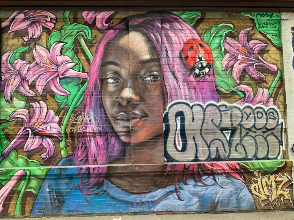 A vivid mural on a city wall features a portrait of a woman with pink-toned hair among stylized, oversized lilies and a ladybug. The artistry displays detailed realism in the subject's face juxtaposed with graphic elements of urban graffiti. The vibrant colors and life-like representation create a captivating street art piece, emblematic of a dynamic urban arts scene.