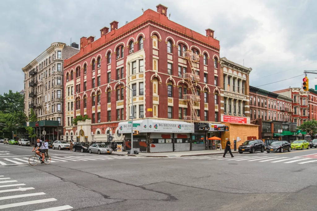 A wide-angle view of a bustling intersection in Harlem, New York City, featuring a cyclist in motion and pedestrians crossing the street, with the striking red and beige facade of a classic building housing a tax preparation service on the corner.