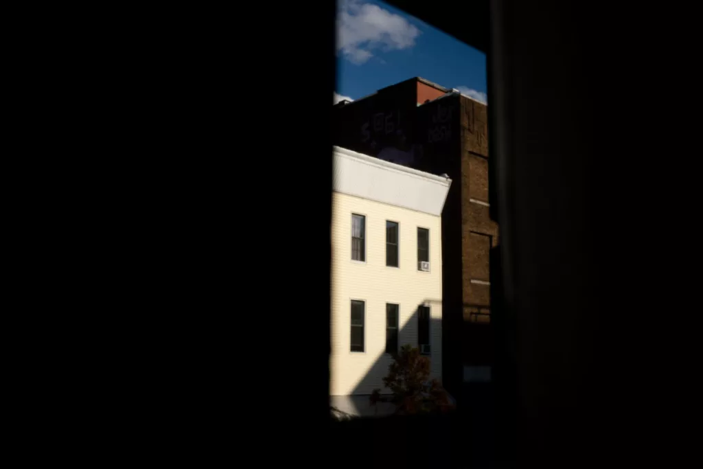 This photo offers a view through a dark opening, possibly a window, that frames a scene of urban architecture. The contrast between the darkness in the foreground and the illuminated buildings creates a natural vignette, focusing the viewer's attention on the well-lit white building with several windows. The light striking the side of the building casts sharp shadows that highlight its texture and form. In the background, there's another building with graffiti on its side, suggesting an urban setting where buildings old and new are layered together, creating a rich visual tapestry. The overall composition has a quiet, contemplative mood, possibly suggesting themes of observation, the passage of time, or the juxtaposition of different elements within a city.