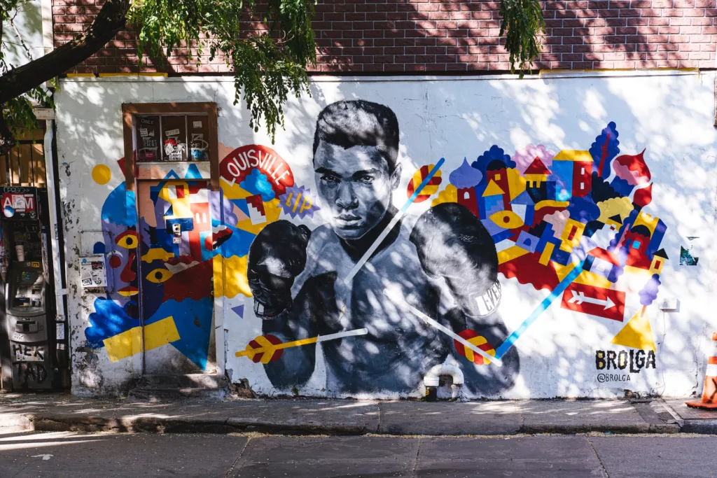 This image features a striking street art mural of Muhammad Ali, the iconic heavyweight boxing champion known as "The Greatest." He is depicted in a classic boxing stance, with gloves raised, against a background filled with colorful abstract shapes and the text "LOUISVILLE LIP," a nod to Ali's hometown and his reputation for verbal sparring. The artwork is vivid and full of movement, highlighting Ali's strong presence both inside and outside the ring. The signature "@BROLGA" credits the artist on the bottom right, indicating this as a piece by the street artist known as Brolga.