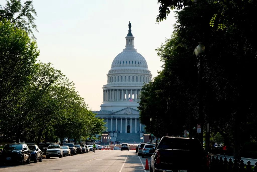 The image depicts a street view leading up to the United States Capitol Building in Washington, D.C. There is a clear sky and the majestic Capitol dome is prominently featured in the background, centered in the composition. The street is lined with parked cars on both sides, and lush green trees flank the sides of the image, adding a natural element to the urban scene. The Capitol's facade is illuminated by the soft light of what appears to be either morning or evening sun, casting gentle shadows and giving the scene a serene atmosphere. Street lamps stand at the edges of the sidewalk, and there are a few indistinct figures near the steps of the Capitol, too small to detail. The image captures the grandeur of the building while also showing a calm, everyday moment on the streets of the capital city.