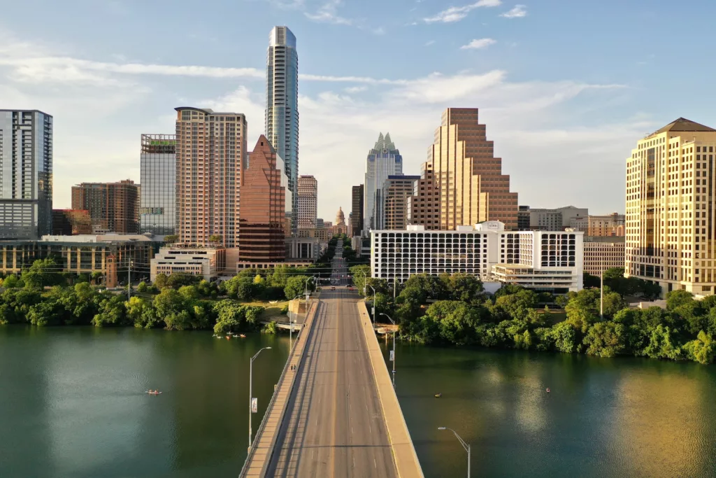 A panoramic view of a Austin skyline featuring modern high-rise buildings with various architectural designs. In the foreground, there is a wide bridge over a calm river with a few small boats. The river is flanked by lush green trees on either side. The sky is clear with a few scattered clouds, suggesting it's a sunny day. The city exudes a mix of urban development and natural beauty.