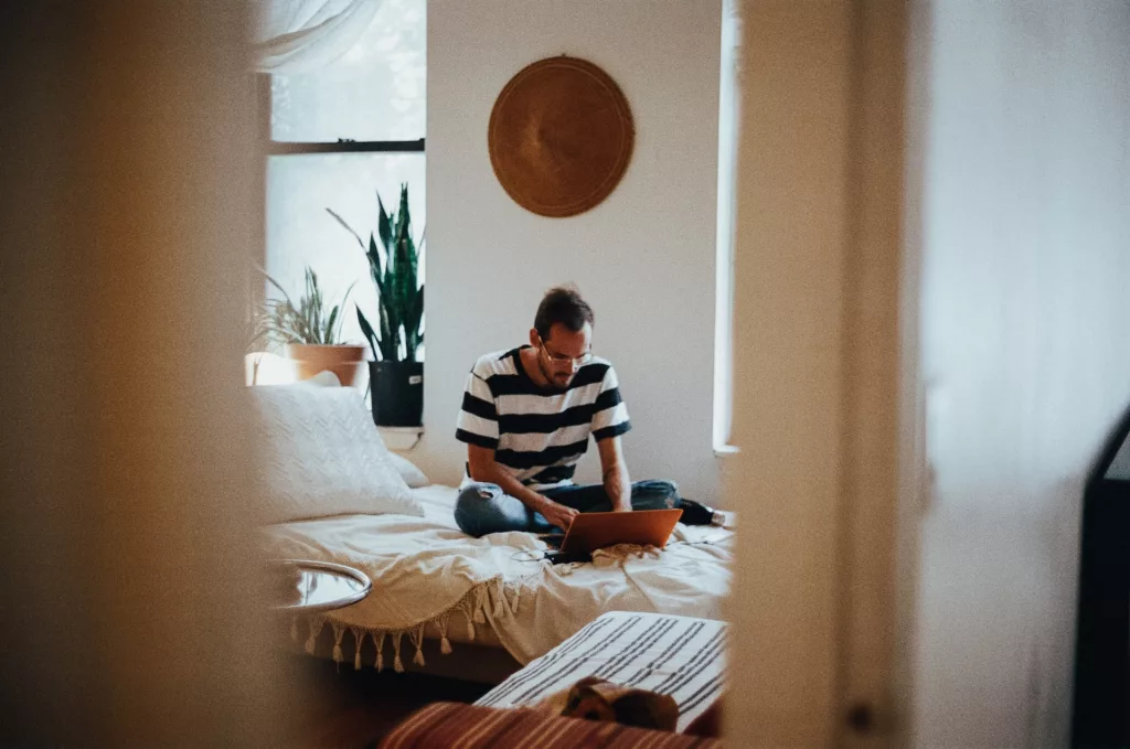 A focused individual benefits from the flexibility of renting by working on a laptop in a serene bedroom space, decorated with plants and minimalistic furnishings, embodying the ease and comfort of a rented home environment.