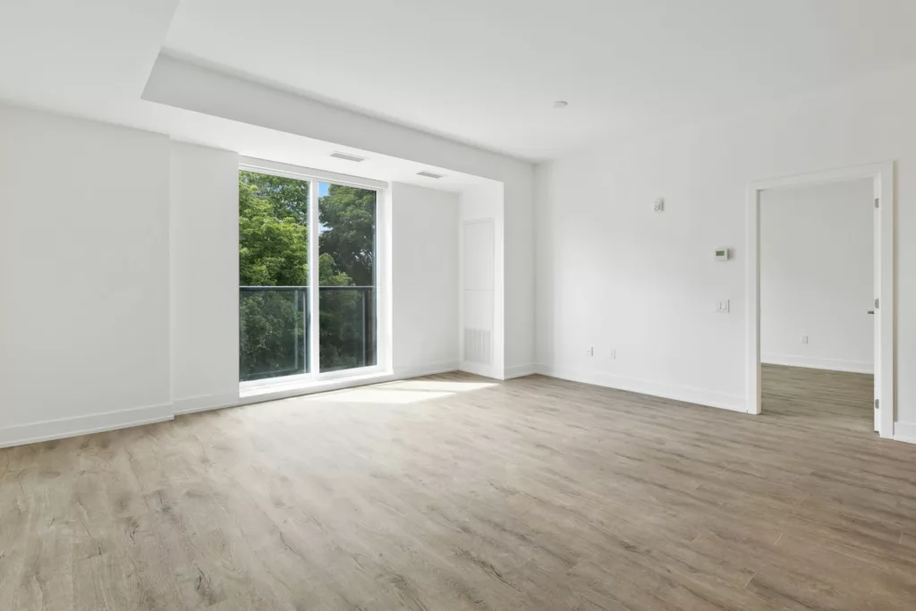 This image features an empty, modern apartment room. The room is bright with natural light coming through a large window with a view of greenery outside. It has white walls, a light wooden floor, and a high ceiling with recessed lighting. On the left, there's a built-in shelf or alcove, and on the right, an open door leads to another room. The space feels clean and new, suggesting it is ready for someone to move in and personalize it.