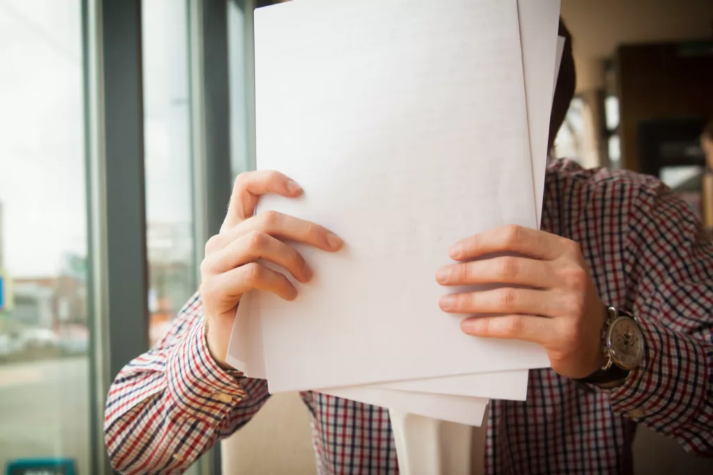 A person in a plaid shirt is holding and reading a stack of papers, obscuring their face. They are standing indoors, next to a window, with natural light illuminating the documents in their hands.