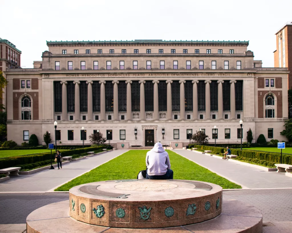 The image shows a person sitting on a circular stone bench in a spacious, open plaza. In front of them is a large, imposing building with classical architectural features, including a series of grand columns and names of classical scholars such as Homer and Aristotle inscribed along its upper facade. This setting is evocative of an academic institution, likely a university, where the architecture aims to inspire scholarly pursuit and reflect a rich intellectual legacy. The well-manicured lawns and clear pathways suggest a well-maintained campus or educational space, which is typical of prestigious universities. The person is facing away from the camera, adding a sense of contemplation and focus, fitting for a college setting where students often engage in study and reflection.