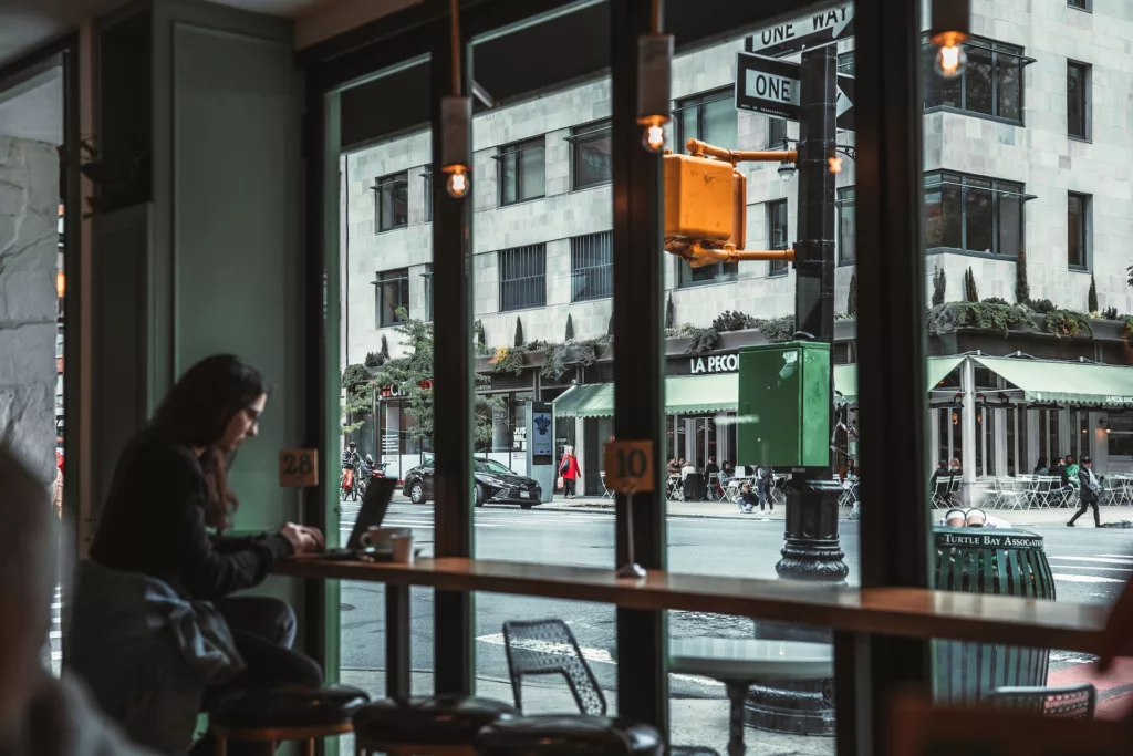 A person sits at a window bar in a cozy cafe, working on a laptop overlooking a bustling New York City street with pedestrians, traffic, and outdoor seating at a restaurant, epitomizing one of the best places to work remotely in NYC.