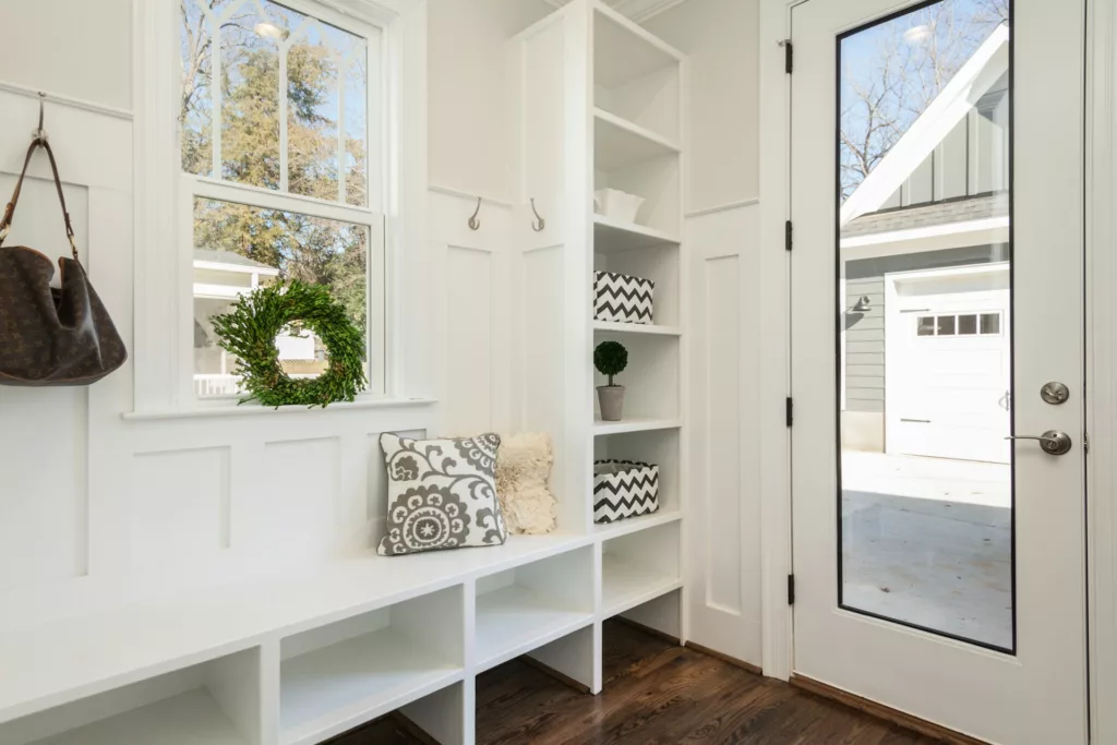 An entryway with a built-in white bench and storage cubbies under a large window adorned with a green wreath. To the right of the bench are shelves with decorative items including a patterned pillow, a small plant, and two storage baskets with chevron patterns. The floor is dark hardwood, and there is a designer handbag hanging from a hook next to the window. The bright space is well-lit, with a view of a garage door through the glass panel of the entry door.
