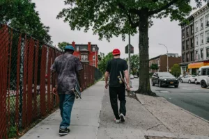 Two individuals casually walking on a sidewalk in Brooklyn, one carrying a skateboard, with the backdrop of historic red brick buildings and urban street scenery, symbolizing the dynamic lifestyle and residential options available when living and renting in Brooklyn.