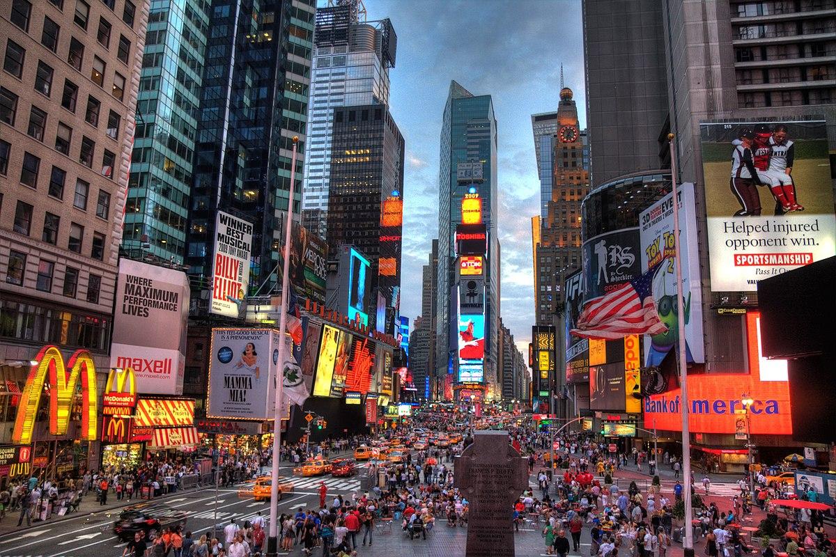 The image depicts a bustling Times Square in New York City at dusk. The sky is a mixture of blue and orange hues, indicating either sunrise or sunset. The area is crowded with people walking or gathering in the pedestrian areas. Numerous bright and colorful billboards and neon signs light up the buildings, advertising various brands, Broadway shows like "Billy Elliot" and "Mamma Mia," and a large digital display showing the countdown for New Year's Eve. Iconic franchises such as McDonald's are visible amidst the commercial splendor. The streets are busy with traffic, including the famous yellow taxis of NYC. A statue or monument stands in the foreground, and the entire scene is a vibrant display of city life and commercial activity, characteristic of one of the most famous urban squares in the world.