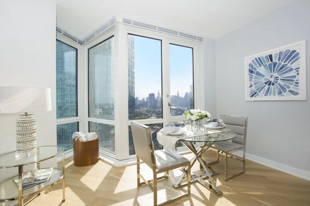 The image features a bright and modern dining area within a high-rise apartment. The room is filled with natural light coming through large corner windows that offer a stunning view of a city skyline. The decor includes a round glass dining table with a reflective gold base, accompanied by four chairs with sleek silver frames and light grey cushioning. A piece of blue and white abstract art hangs on the wall, complementing the room's clean, contemporary aesthetic. A stylish lamp sits on a glass side table, and a brown leather container holds decorative items. The floor is adorned with light hardwood, and the overall ambiance of the room is elegant and airy.