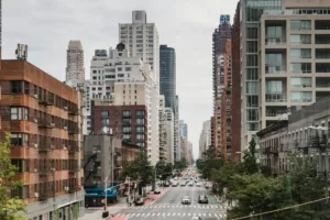 Urban street view of one of New York City's safe neighborhoods, showcasing a mix of modern and traditional architecture with residential and commercial buildings lining the road. The street is bustling with cars and the occasional bus, highlighting the area's livability and the presence of green trees suggests a pleasant, community-friendly environment.