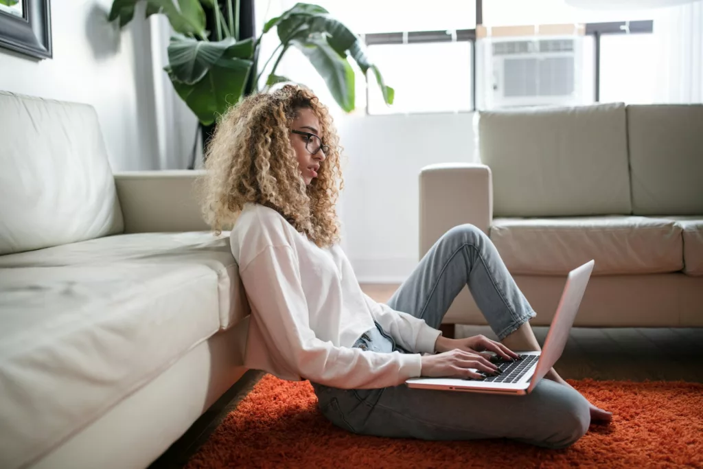 A young woman with curly hair and glasses sits on a floor with a laptop, focused on the screen. She is casually dressed in a light-colored top and gray sweatpants, suggesting a relaxed home environment. The room is well-lit with natural light and furnished with modern decor, including a comfortable sofa and indoor plants, creating an atmosphere of a cozy living space.
