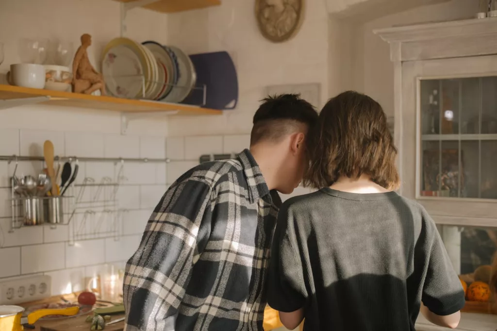 Two people stand side by side in a cozy kitchen, facing away from the camera, suggesting a scene of domestic collaboration. One with a short, undercut hairstyle dressed in a plaid shirt, and the other with shoulder-length hair in a dark grey shirt, seem to be jointly engaged in a task, perhaps preparing a meal or considering an item on the counter. The kitchen exudes warmth, adorned with hanging utensils and a variety of dishes on open shelves. This setting complements the concept discussed in "The Roommate Chore Chart They Absolutely Can't Ignore" article, illustrating the essence of shared duties and the importance of organization and teamwork in creating a harmonious living space.
