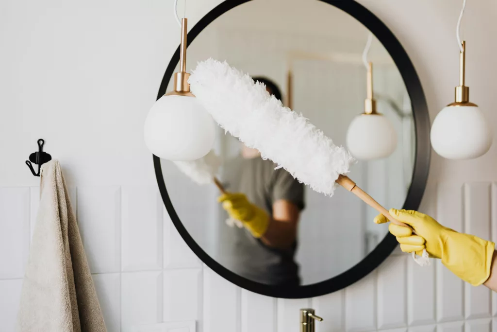 A person wearing yellow gloves is cleaning a mirror with a white, fluffy duster in a bathroom. The mirror reflects the image of the duster, enhancing the focus on the cleaning activity. This image could accompany an article with tips for keeping a rented apartment clean and organized, illustrating the importance of regular maintenance like mirror cleaning.