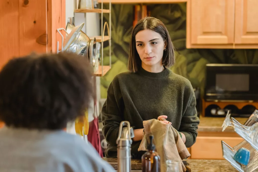 A young woman in a dark green sweater stands in a kitchen, engaged in a conversation with an unseen person. Her focused expression and relaxed posture suggest a casual and interactive environment. The kitchen features wooden cabinets and a variety of everyday items such as a tea kettle, glasses, and bottles, indicating a shared living space. This image is well-suited for an article discussing the pros and cons of renting and living in shared apartments.