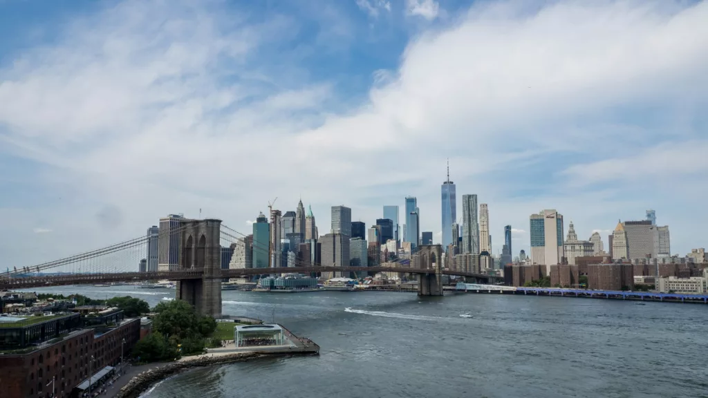 This is an image of the New York City skyline viewed from Brooklyn. The Brooklyn Bridge stretches across the left side of the frame, connecting to the dense collection of skyscrapers of Lower Manhattan, including the prominent One World Trade Center. The East River flows in the foreground, with boats navigating the water and a riverside park visible on the bottom left. The sky is partly cloudy, casting soft light over the urban landscape.