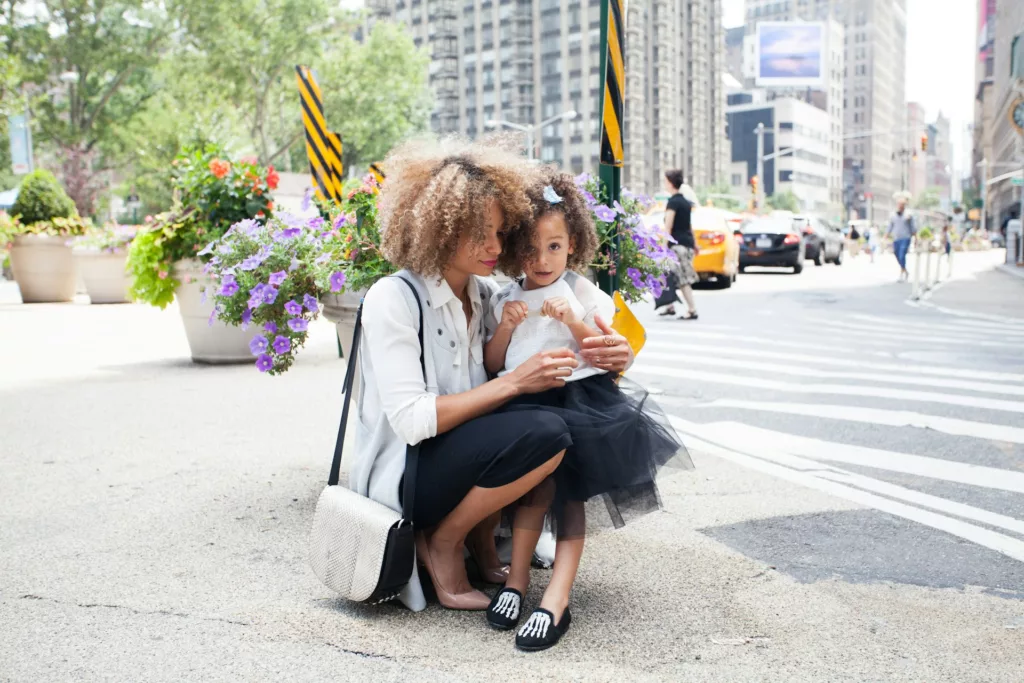 A mother with curly hair is crouching on a city sidewalk, holding her young daughter who is dressed in a black tutu and white shirt. They are surrounded by large flower pots with colorful blooms, and city buildings and yellow taxis are visible in the background. Both are looking towards the camera with the mother smiling gently and the daughter with a curious expression. This image illustrates how flexible leases can ease your move to New York City, allowing families to settle in quickly and comfortably in their new urban environment.