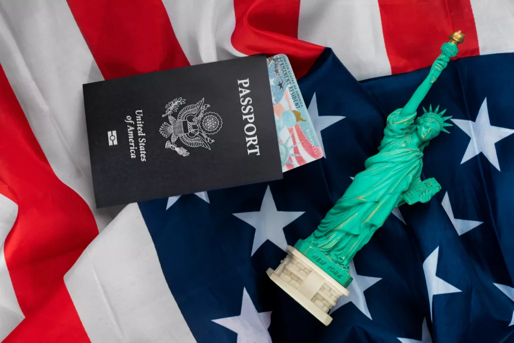An image of a U.S. passport with a visa tucked inside, placed on an American flag. Next to the passport is a small statue of the Statue of Liberty.