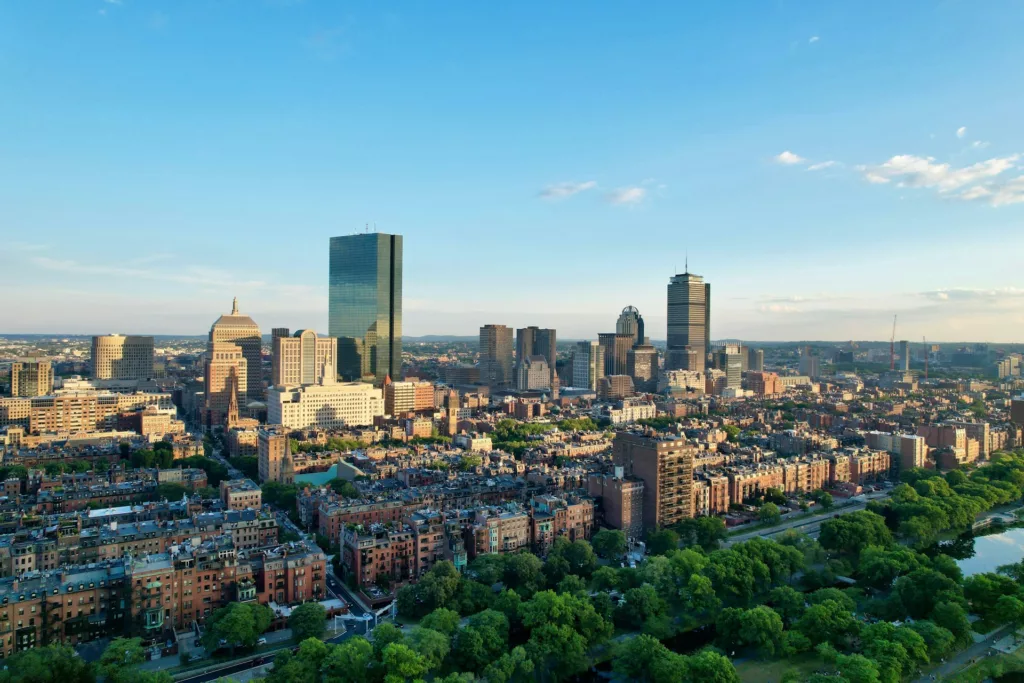 Aerial view of Boston showcasing the city's skyline with high-rise buildings and lush green parks, highlighting a mix of historic and modern architecture under a clear blue sky.