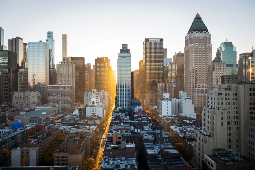 A sunrise view of a bustling cityscape featuring a mix of tall modern skyscrapers and older buildings. The sun's rays are peeking through the buildings, casting a warm glow on the high-rise structures and the streets below.