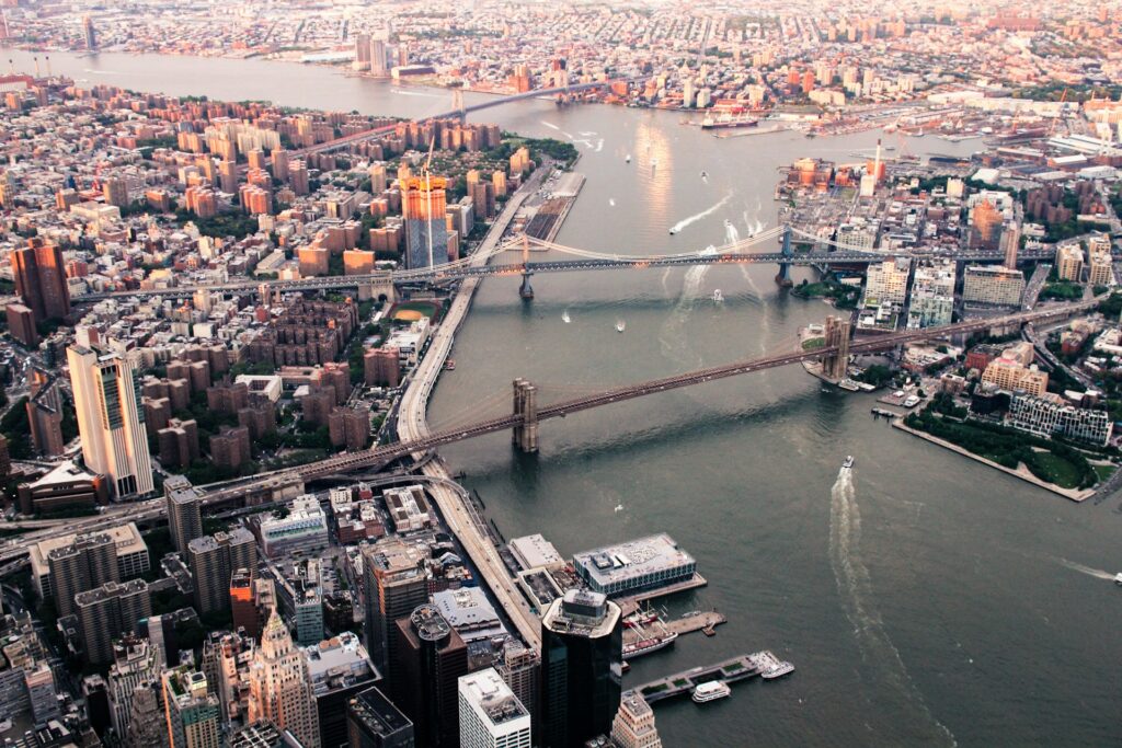 Aerial view of New York City, featuring the Brooklyn Bridge and the Manhattan Bridge spanning the East River. The image showcases the densely packed cityscape of Lower Manhattan, with various skyscrapers, buildings, and streets visible. The river is dotted with boats, and the horizon shows the sprawling urban landscape of Brooklyn.