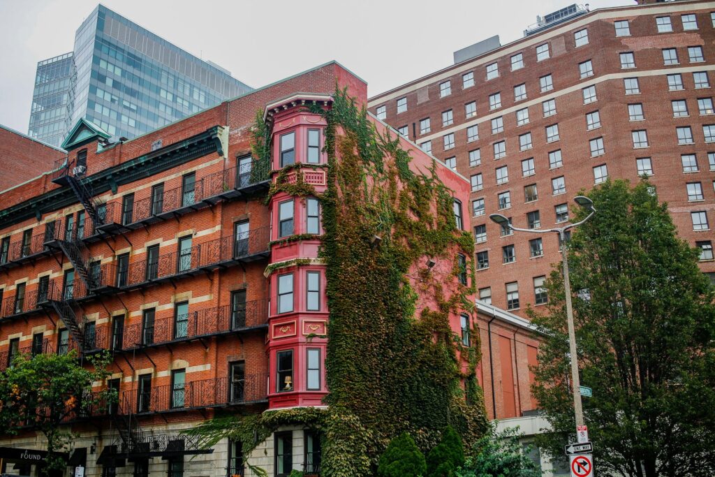 A cityscape featuring a historic red Boston brick building partially covered in ivy, with an ornate red and gold bay window section. The building has classic fire escapes. Modern high-rise buildings and trees surround the historic structure.