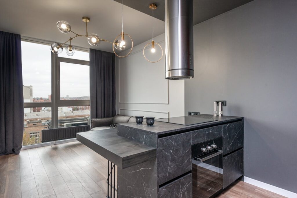 Modern kitchen with sleek black marble countertops, an integrated stove, and a large metallic range hood. The space features contemporary pendant lighting, floor-to-ceiling windows with dark curtains, and a wooden floor. The kitchen island has a unique, asymmetrical design with a built-in seating area, and the room offers a view of the cityscape outside.