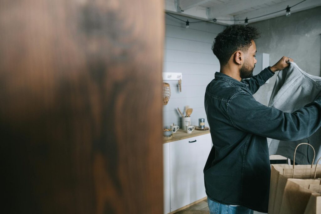 A man with short curly hair and a beard is standing in a modern kitchen. He is holding up a gray shirt, possibly examining or folding it. In the background, there are kitchen utensils and shelves with various items. The overall scene gives a cozy, homey feel.