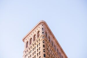 The image shows the top portion of a tall, iconic building with a flatiron shape, characterized by its narrow, triangular design. The building features ornate details on its facade, with rows of windows and a light stone exterior. The background is a clear, light blue sky. The angle of the photograph emphasizes the unique, pointed architecture against the open sky.