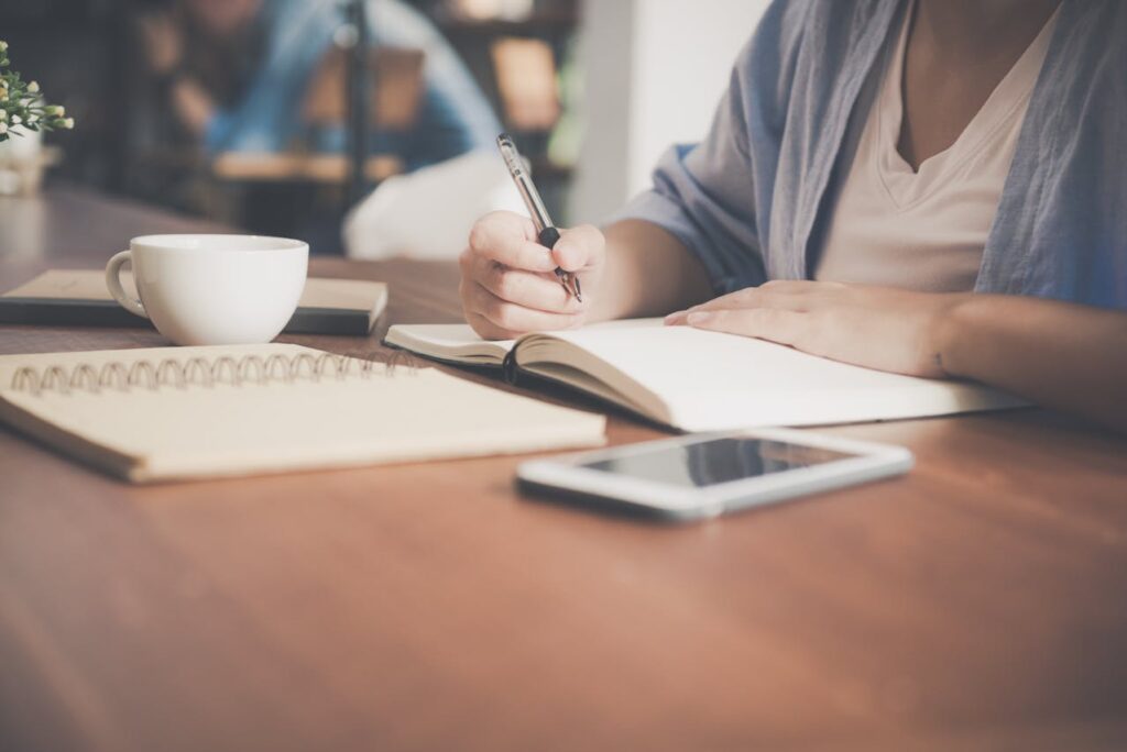 A woman writing on a notebook beside a teacup and tablet computer.