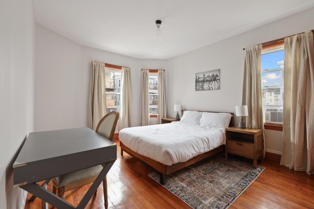 Bright bedroom with wooden floors featuring a large bed with white linens, a desk and chair, two bedside tables with lamps, and three windows with light curtains. The room for rent is minimally decorated with a small wall art piece above the bed, creating a clean and airy atmosphere.