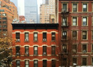 The image shows several brick apartment buildings in an urban setting. The buildings have a classic red-brick façade with fire escapes and air conditioning units in the windows. In the background, there are taller, more modern skyscrapers, creating a contrast between the older and newer architecture. The trees in front of the buildings appear leafless, indicating a colder season, possibly late fall or winter.