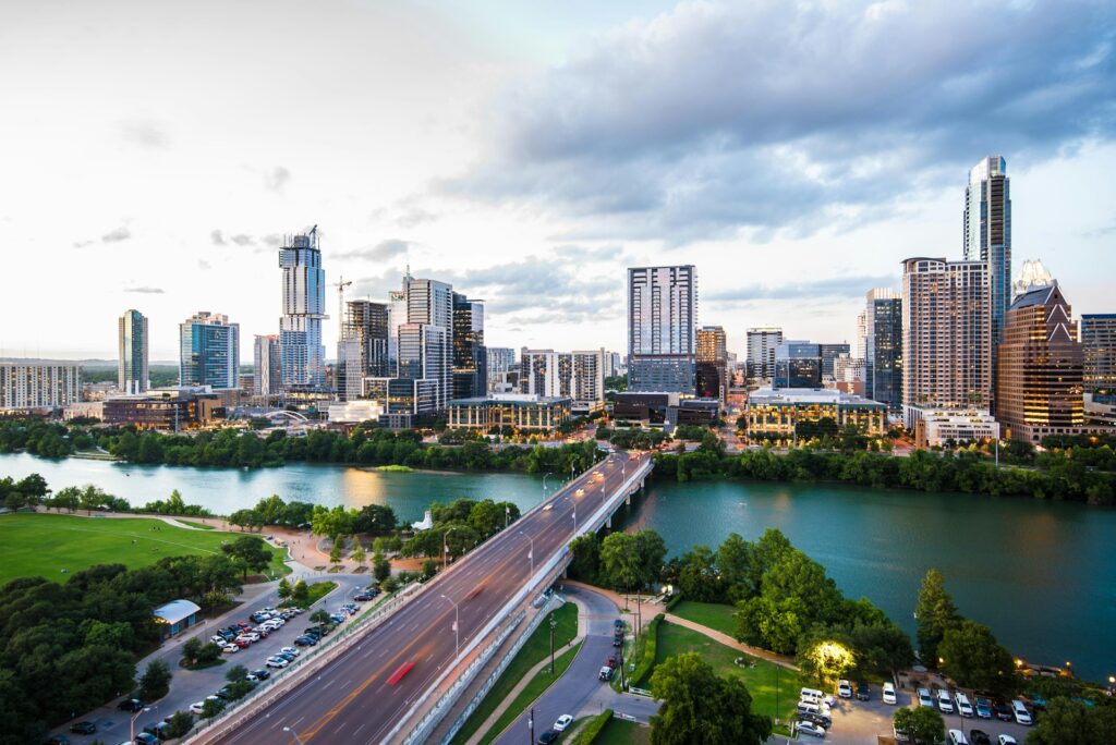 A scenic view of Austin, Texas skyline with a river and bridge in the foreground, under a partly cloudy sky. Article title: 'Best 13 Neighborhoods In Austin To Live And Rent.