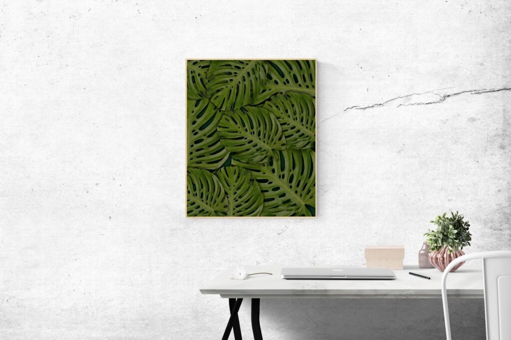 Framed tropical leaf print on a minimalist office wall, with a clean white desk, laptop, and potted plant for a modern, nature-inspired workspace.